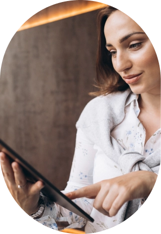 Kobieta w jasnej bluzce i szarym swetrze uśmiecha się, korzystając z tabletu. Stoi przed nowoczesną ścianą w delikatnym, ciepłym świetle. Panuje atmosfera skupienia i radości.